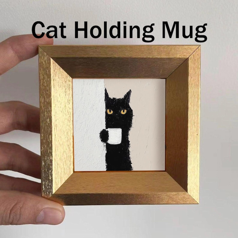 May include: A framed artwork featuring a black cat holding a white mug. The cat has yellow eyes and is set against a neutral background. The frame is a gold color, and the text "Cat Holding Mug" is at the top.