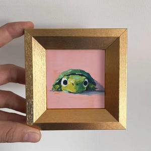 May include: A small, square painting of a green turtle peeking out from a pink background. The turtle has large, round eyes and a yellow face. The artwork is framed in a gold-colored wooden frame.