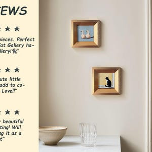 May include: Two framed cat paintings hang on a cream-colored wall. One painting depicts two white birds on a blue background. The other shows a black cat on a yellow background. Both are in square, gold-toned frames.