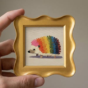 May include: A small square painting of a hedgehog with rainbow-colored quills, featuring a pink head and a cream-colored face, encased in a decorative gold-toned frame with a wavy, scalloped edge and a smooth, matte finish.