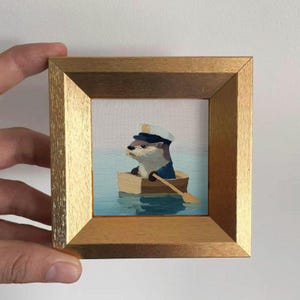 Könnte beinhalten: Gerahmtes Gemälde, das einen Otter in einem kleinen Holzboot zeigt, der eine Kapitänsmütze und Uniform trägt. Das Kunstwerk zeigt einen ruhigen blauen Wasserhintergrund und einen goldfarbenen Rahmen, der ein skurriles nautisches Thema schafft.
