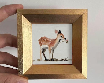 May include: A framed miniature painting of a fawn holding a pink flower in its mouth. The fawn has brown and white spots and is set against a white background. The frame is gold-colored.