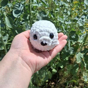 Könnte beinhalten: Ein kleines, weißes, gehäkeltes Seehund-Plüschtier mit schwarzen Augen und einem schwarzen Mund. Der Seehund wird in einer Hand vor einem Hintergrund aus grünem Laub und gelben Blumen gehalten. Das Plüschtier hat ein strukturiertes, flauschiges Aussehen.