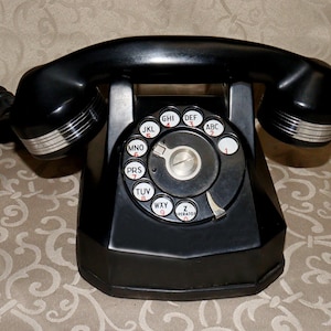 May include: A black rotary telephone featuring a handset with silver-toned ribbed accents on the ends. The base includes a circular dial with white buttons displaying black letters and red numbers, connected by a black coiled cord to the main unit.