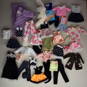 May include: A selection of doll clothing, including dresses, skirts, trousers, and jackets. The garments feature various patterns, such as polka dots, floral prints, and gingham. Colours include pink, black, white, and blue. Some outfits have bows and lace trim.