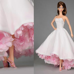 Könnte beinhalten: Ein weißes Kleid mit einem rosa, gerüschten Rock. Das Kleid ist trägerlos und hat ein tailliertes Oberteil. Die Puppe trägt rosa High Heels.
