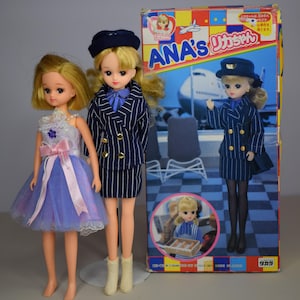 May include: Two blonde-haired dolls, one in a blue and white striped suit and the other in a blue dress with a pink bow. A box with the text "ANA's" and Japanese text features a doll in a similar outfit.