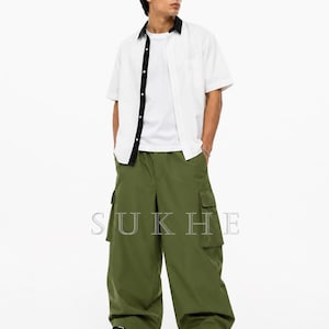 Könnte beinhalten: Ein weißes Kurzarmhemd mit schwarzem Kragen und Besatz, getragen über einem weißen T-Shirt. Das Outfit umfasst olivgrüne Cargohosen und schwarz-weiße Turnschuhe. Das Wort "SUKHE" ist auf die Hose gedruckt.