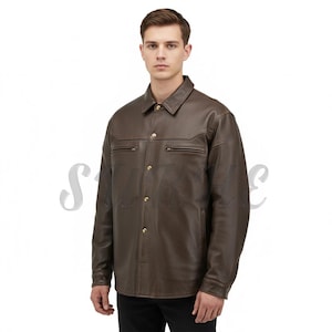 May include: A brown leather jacket with a collared neckline and button-down closure. The jacket features two chest pockets with zippers and gold-coloured buttons. The model is wearing black trousers.