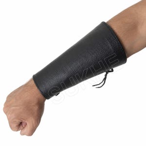May include: A black leather forearm bracer featuring a textured surface and a lace-up closure on the side. The protective gear is designed to fit snugly around the lower arm, with visible stitching along the edges and thin black cord ties.