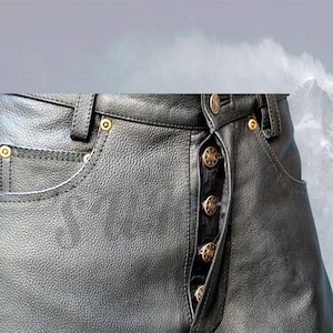 Puede incluir: Primer plano de pantalones de cuero negro con detalles de botones dorados. Los pantalones presentan un diseño clásico de cinco bolsillos con bragueta abotonada. El cuero tiene un aspecto texturizado y las costuras son visibles.