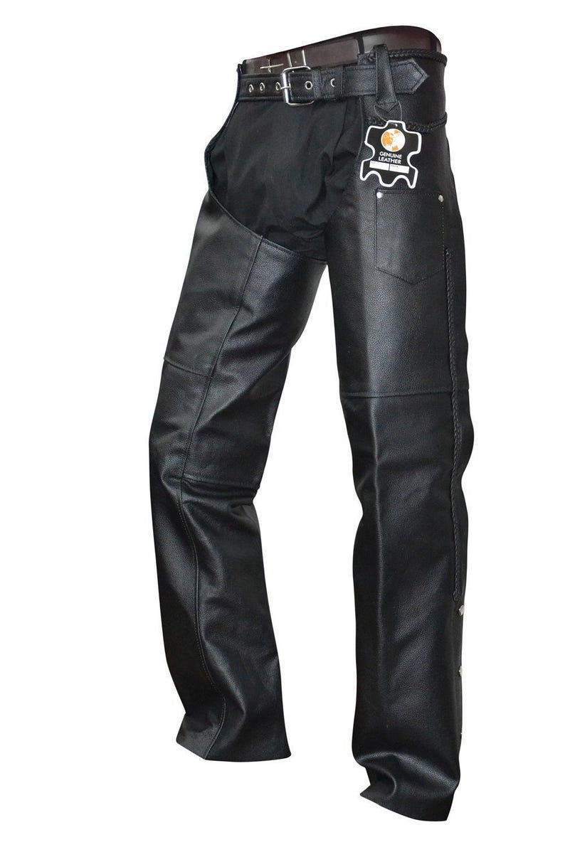 May include: Black leather motorcycle chaps with a belt and buckle closure. The chaps have a classic design with a pocket on the right leg and decorative stitching. A tag that says "Genuine Leather" is attached.
