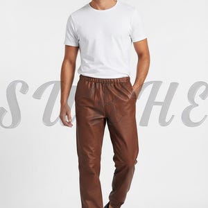 May include: A person wearing a white crew neck t-shirt and brown leather pants. The pants have an elastic waistband and are cuffed at the ankles. The person is also wearing black sneakers.