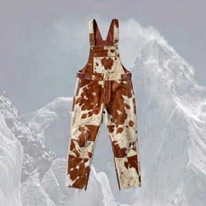 May include: Overalls with a cowhide pattern in shades of brown and white. The overalls have adjustable straps, pockets, and zippers on the legs. The bib and straps are a solid brown color.