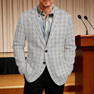 May include: A light grey blazer patterned with black anchors. The blazer has a two-button closure and is worn over a denim shirt. The person is wearing black trousers. The background includes a stage and a wooden podium.