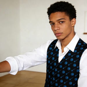 May include: A black vest with a blue pattern, worn over a white collared shirt. The vest features a repeating design of a blue key-like symbol. The image shows a person wearing the vest.