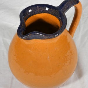 May include: A ceramic pitcher with a rounded body and a handle. The pitcher is a warm orange color with a dark blue rim and handle accents. The interior is also orange. This handmade pitcher is suitable for serving beverages.