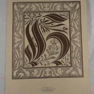 May include: A framed print featuring a stylized letter "S" in a brown, floral design. The letter is surrounded by an ornate border with a repeating pattern. The background is a light beige color. The print is a vintage piece.