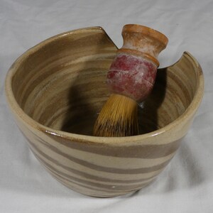 May include: A ceramic shaving bowl with a brown and tan striped design. A shaving brush with a red handle and yellow bristles rests inside the bowl. The bowl has a unique curved shape.