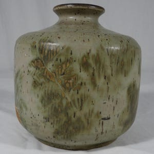May include: A cylindrical ceramic vase with a narrow neck and a wide, rounded body. The surface features a speckled beige and sage green glaze with dark brown vertical streaks and a faint, leaf-like botanical imprint pattern across the exterior.