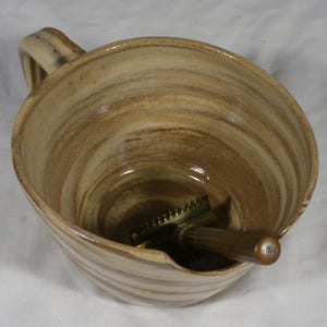 May include: A beige and brown swirl-patterned ceramic shaving mug with a single handle and a notched rim. Inside the mug rests a safety razor featuring a metallic gold-toned head and a dark brown wooden handle, positioned at an angle.