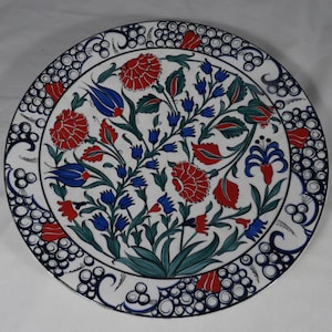 May include: A round, white ceramic plate with a floral design. The plate features red, blue, and green flowers and leaves. The border is white with a blue pattern. The plate is a decorative item.