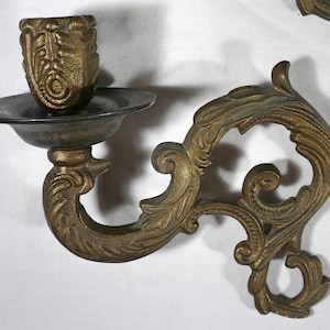 May include: A wall-mounted candle holder crafted from aged brass, featuring an ornate, scrolling metal arm with intricate leaf-like patterns. The piece includes a circular drip tray and a textured, cylindrical candle cup for holding a taper.