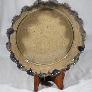 May include: A handmade ceramic pie plate with a scalloped edge. The plate has a speckled tan interior and a dark blue-grey rim. It is displayed on a wooden stand, showcasing its rustic charm and artisanal quality.