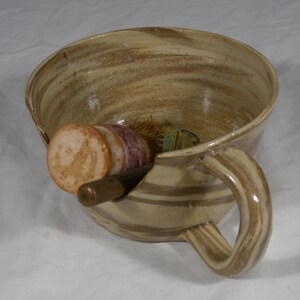 May include: A beige and brown marbled ceramic shaving bowl with a side handle and a notched rim. Inside the bowl rests a shaving brush with a light-colored wooden handle and a dark brown handle tool, both positioned within the bowl's interior.