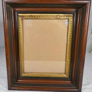 May include: A rectangular picture frame featuring a wide, dark brown wooden outer border with a stepped profile. The inner edge is accented by a thin black border and a gold-toned trim decorated with a repeating embossed pattern surrounding the glass.