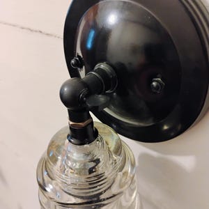 May include: A black wall sconce with a clear, ribbed glass shade. The sconce features a circular backplate and a curved arm connecting to the glass shade. The fixture is designed for wall mounting and has a vintage aesthetic.