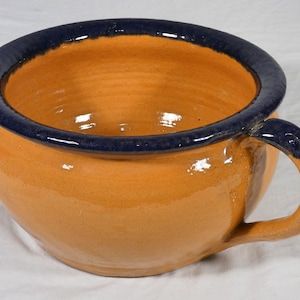 May include: A handmade ceramic bowl with a handle. The bowl is a warm orange color with a dark blue rim and handle accents. The bowl is round and has a wide opening, perfect for serving soups or stews. The bowl measures approximately 6 inches in diameter.