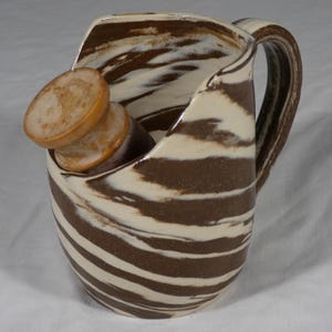 May include: A ceramic pitcher featuring a marbled pattern of dark brown and cream swirls, equipped with a curved handle on the side. A cylindrical wooden pestle with a light tan, textured top rests inside the pitcher, angled toward the rim.