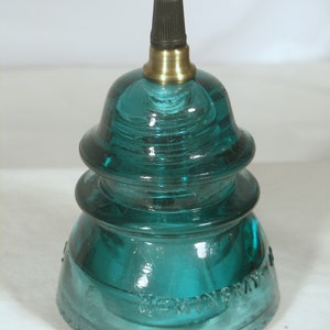 May include: A teal blue glass insulator with a brass top and black cord. The insulator has two tiers and the bottom tier has the text "HEWITT GRAY" embossed on it.