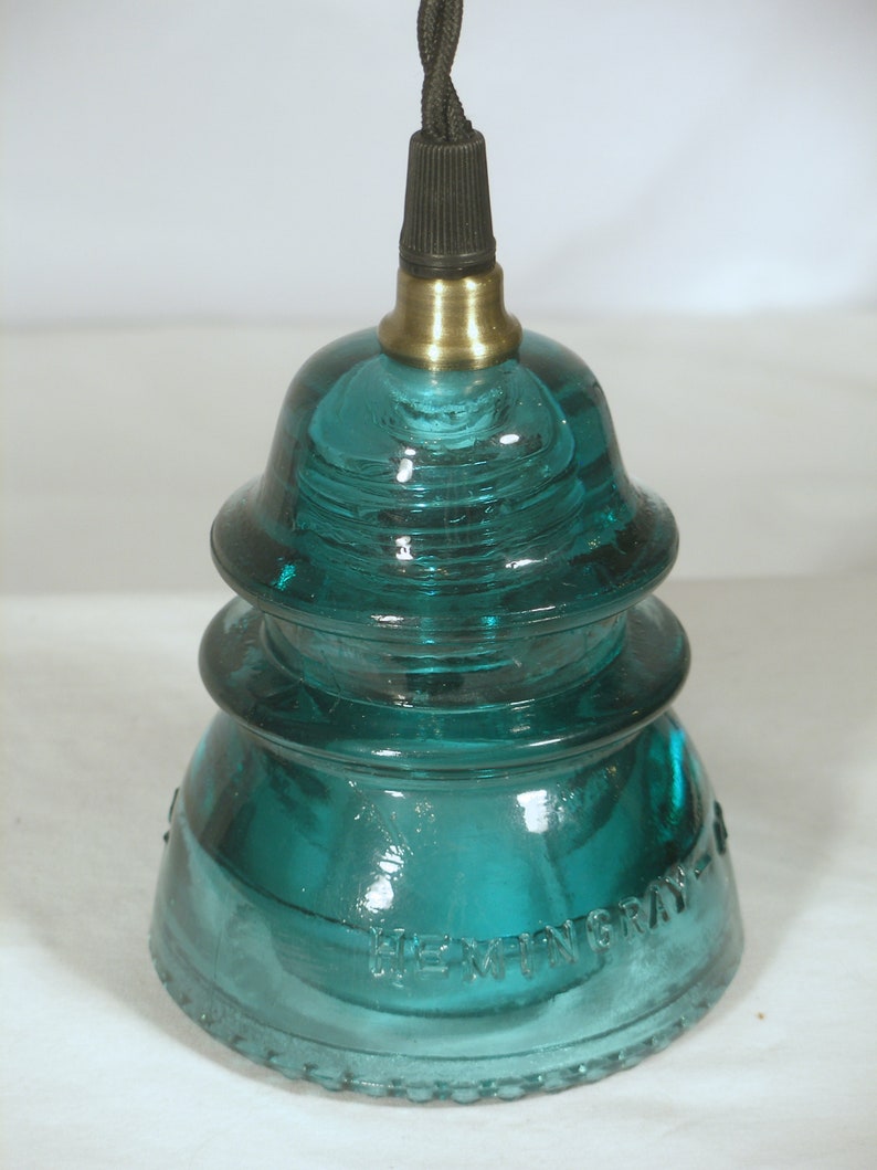 May include: A teal blue glass insulator with a brass top and black cord. The insulator has two tiers and the bottom tier has the text "HEWITT GRAY" embossed on it.