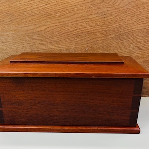 May include: A rectangular wooden box in a deep reddish-brown hue. The box features a flat top with a raised lid and is crafted with visible wood grain. The design is simple and elegant, suitable for storage or display, measuring approximately 30 cm in length.