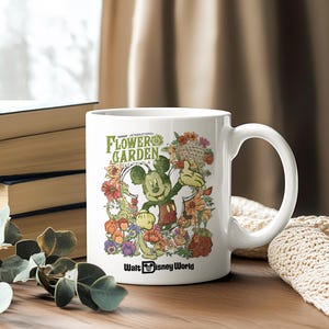 May include: White ceramic mug featuring Mickey Mouse surrounded by colourful flowers. The mug has the text "EPCOT INTERNATIONAL FLOWER GARDEN" and "Walt Disney World". The mug has a large handle.