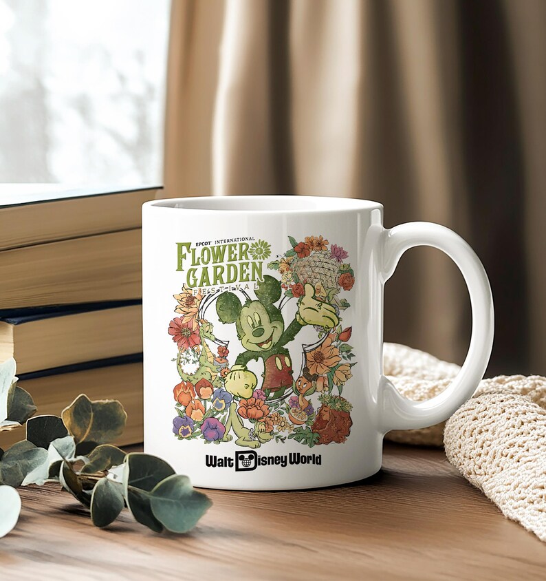 May include: White ceramic mug featuring Mickey Mouse surrounded by colourful flowers. The mug has the text "EPCOT INTERNATIONAL FLOWER GARDEN" and "Walt Disney World". The mug has a large handle.