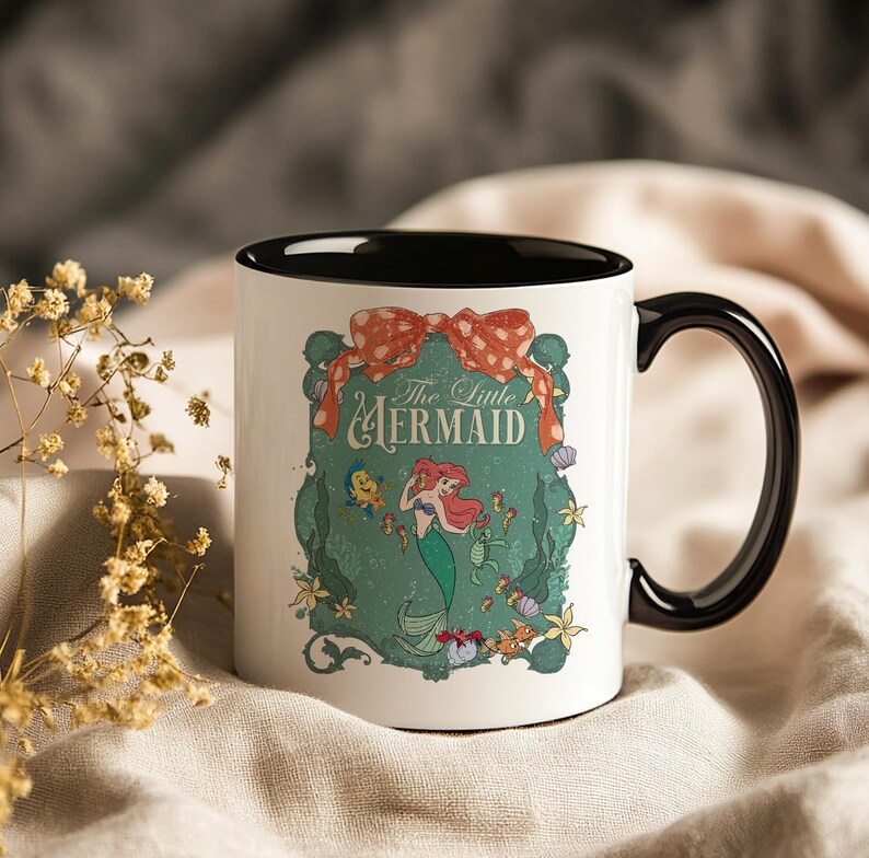 Pode incluir: Caneca de cer&acirc;mica branca com al&ccedil;a e interior pretos. A caneca apresenta uma ilustra&ccedil;&atilde;o colorida de Ariel de A Pequena Sereia, com o texto "The Little Mermaid" em uma fonte decorativa. O design inclui um la&ccedil;o vermelho e v&aacute;rias criaturas marinhas.