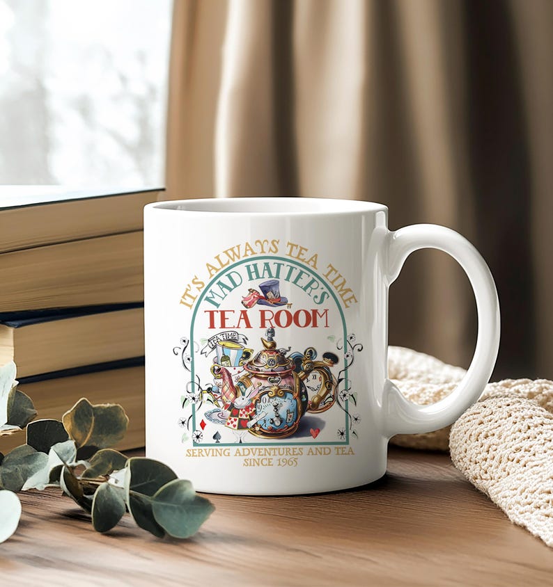 Puede incluir: Taza de cer&aacute;mica blanca con una ilustraci&oacute;n colorida de una tetera, un reloj y naipes. La taza tiene el texto "It's Always Tea Time, Mad Hatter's Tea Room, Serving Adventures and Tea Since 1965".
