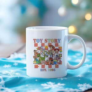 May include: White ceramic mug with a colorful illustration of the Toy Story characters, including Woody, Buzz Lightyear, and Jessie. The text "TOY STORY EST. 1995" is printed on the mug.