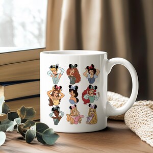 May include: White ceramic mug featuring cartoon illustrations of various princesses wearing mouse ears. The princesses are depicted in a colorful and whimsical style, with each character's unique features and outfits. The mug has a large handle.