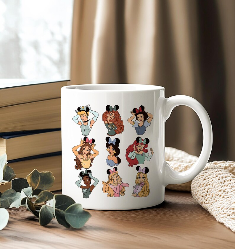May include: White ceramic mug featuring cartoon illustrations of various princesses wearing mouse ears. The princesses are depicted in a colorful and whimsical style, with each character's unique features and outfits. The mug has a large handle.