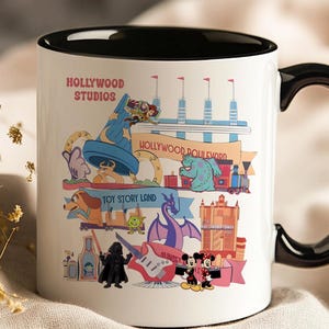 K&ouml;nnte beinhalten: Wei&szlig;er Keramikbecher mit schwarzem Innenraum und Griff, mit farbenfrohen Illustrationen von Hollywood Studios Attraktionen und Charakteren. Der Becher enth&auml;lt die Texte "Hollywood Studios" und "Hollywood Boulevard".