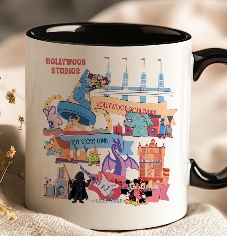 K&ouml;nnte beinhalten: Wei&szlig;er Keramikbecher mit schwarzem Innenraum und Griff, mit farbenfrohen Illustrationen von Hollywood Studios Attraktionen und Charakteren. Der Becher enth&auml;lt die Texte "Hollywood Studios" und "Hollywood Boulevard".