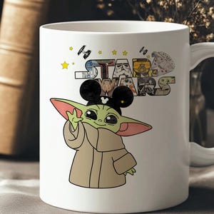 Może przedstawiać: Biały ceramiczny kubek z kresk&oacute;wkową ilustracją Baby Yody w uszach Myszki Miki, wraz z logo Gwiezdnych Wojen i r&oacute;żnymi postaciami. Kubek ma uchwyt i jest ozdobiony gwiazdami i statkami kosmicznymi.