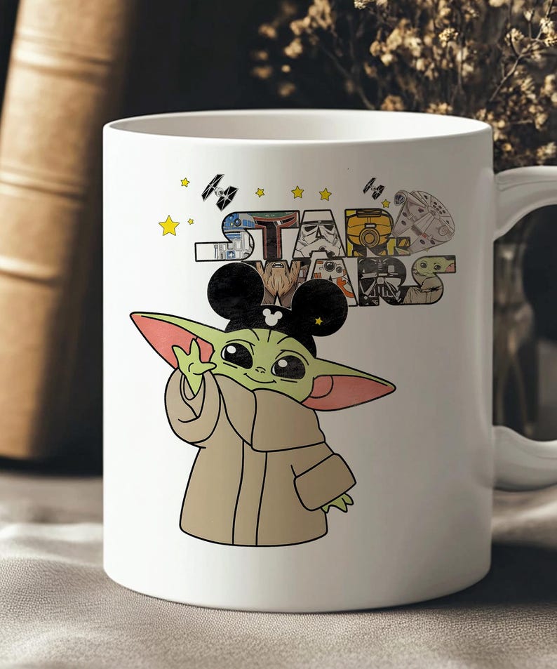 Może przedstawiać: Biały ceramiczny kubek z kresk&oacute;wkową ilustracją Baby Yody w uszach Myszki Miki, wraz z logo Gwiezdnych Wojen i r&oacute;żnymi postaciami. Kubek ma uchwyt i jest ozdobiony gwiazdami i statkami kosmicznymi.