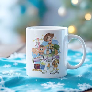 May include: White ceramic mug with a colourful illustration of characters from the animated film Toy Story. The design includes Woody, Buzz Lightyear, Rex, and other characters. The mug has a curved handle and is set against a blurred background.