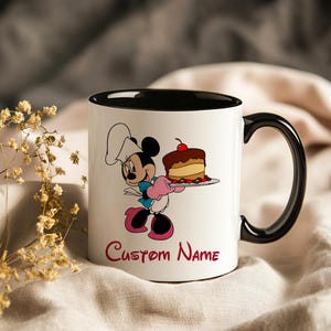 Pu&ograve; includere: Tazza in ceramica bianca con interno e manico neri. La tazza presenta un'immagine a cartoni animati di Minnie Mouse nei panni di uno chef, che tiene in mano una torta. Le parole "Custom Name" sono scritte in rosso sotto l'immagine.