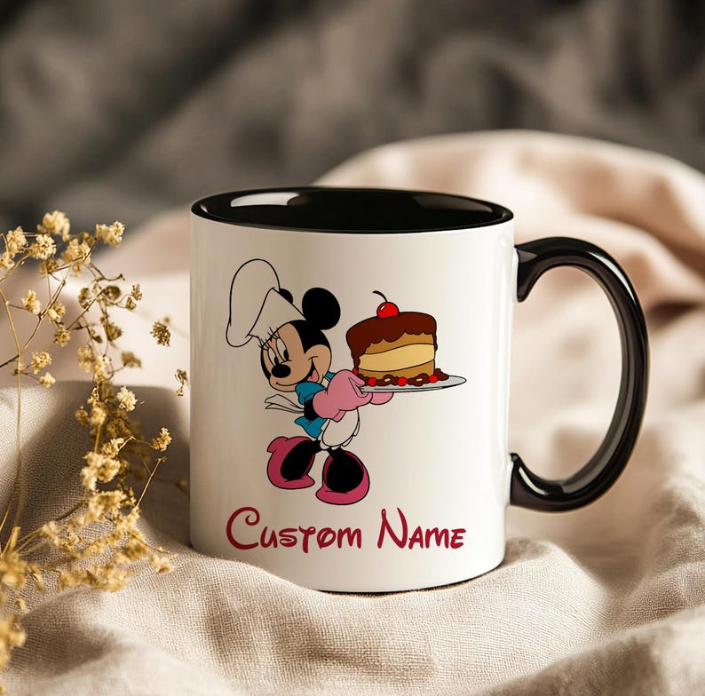 Pu&ograve; includere: Tazza in ceramica bianca con interno e manico neri. La tazza presenta un'immagine a cartoni animati di Minnie Mouse nei panni di uno chef, che tiene in mano una torta. Le parole "Custom Name" sono scritte in rosso sotto l'immagine.